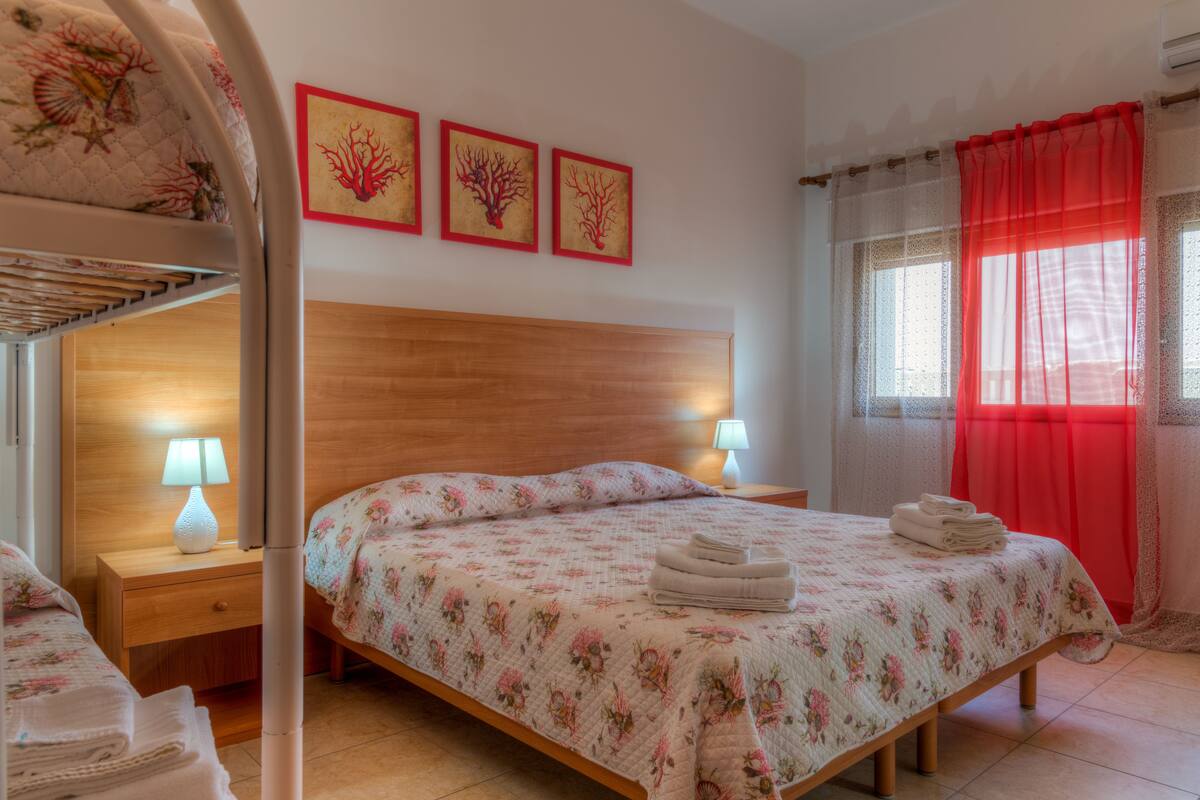 A bedroom with a wooden headboard, floral bedding, a bunk bed, and a window with red curtains.
