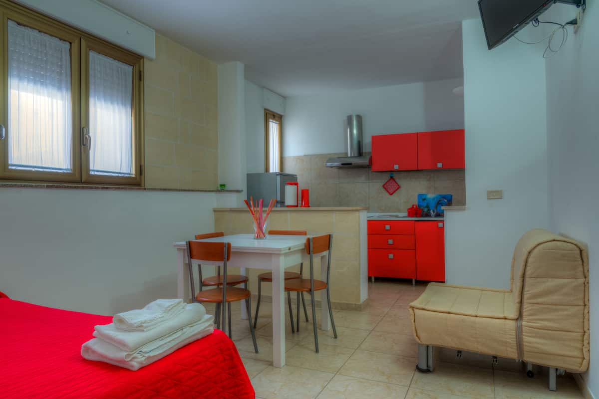 A compact room with a bed, a small dining table with chairs, a kitchenette, and a single chair.