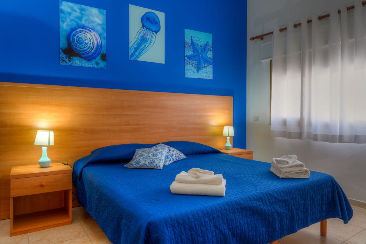 A hotel room with a blue bedspread, two nightstands with lamps, a wooden headboard, and artwork on the wall.