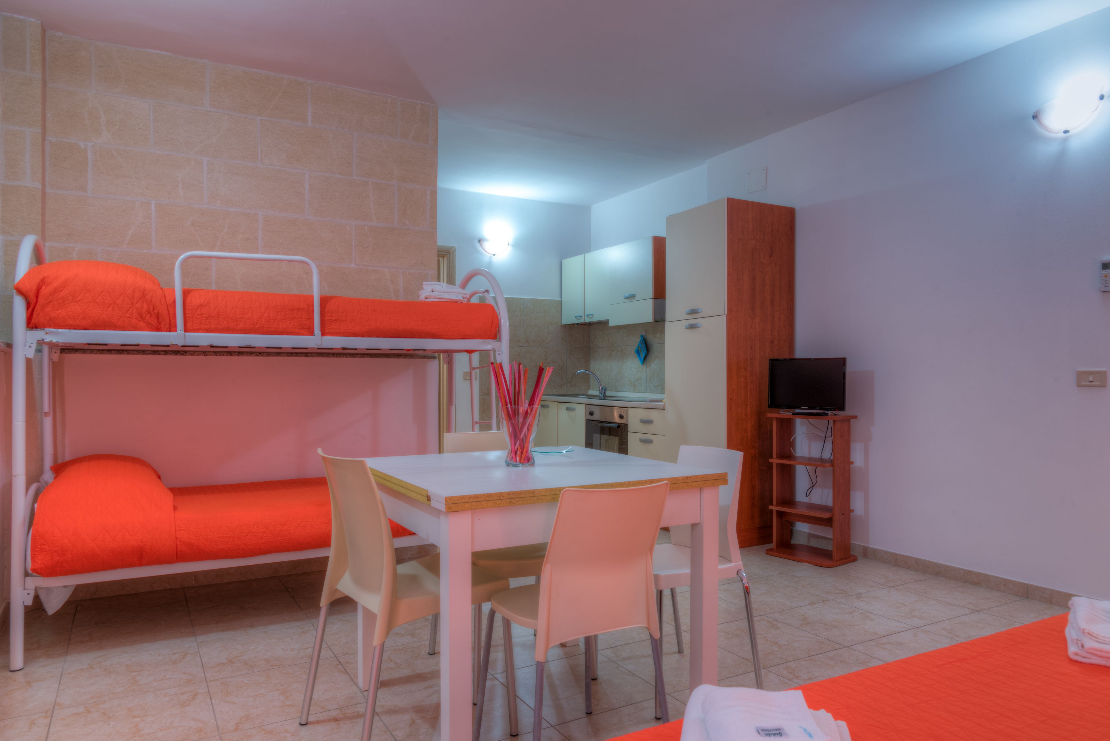 Una cama litera con ropa de cama naranja, una pequeña cocina con fregadero y armarios, un televisor sobre un soporte de madera y una mesa de comedor con sillas.