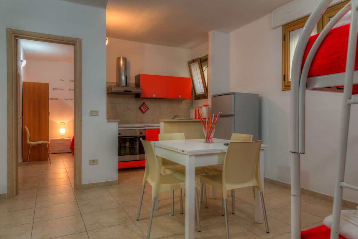 A compact kitchen with a dining area, including a table with chairs, a red cabinet, and a bunk bed with a red mattress.
