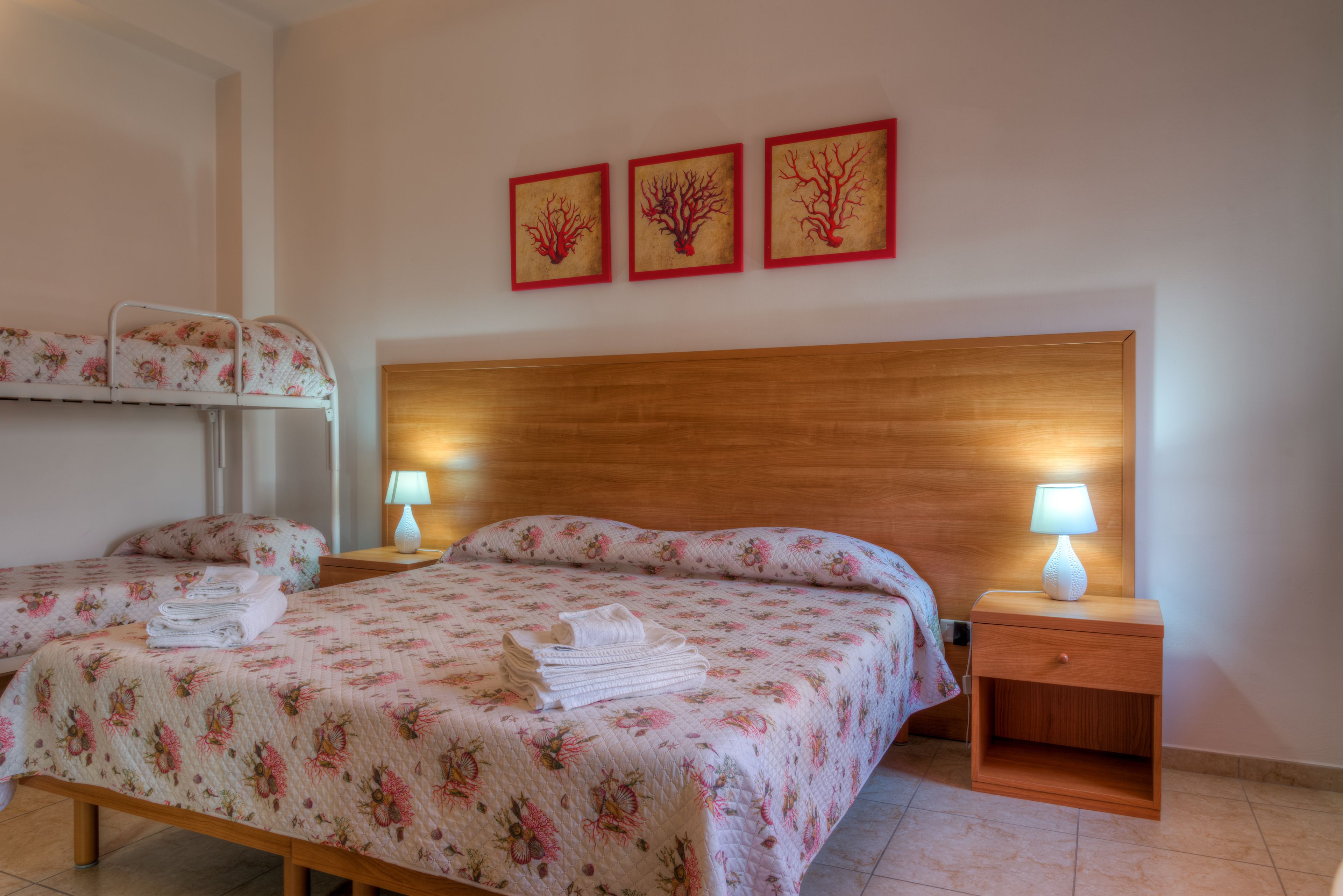 A hotel room with a double bed, two single beds, a wooden headboard, bedside lamps, and three framed wall art pieces.
