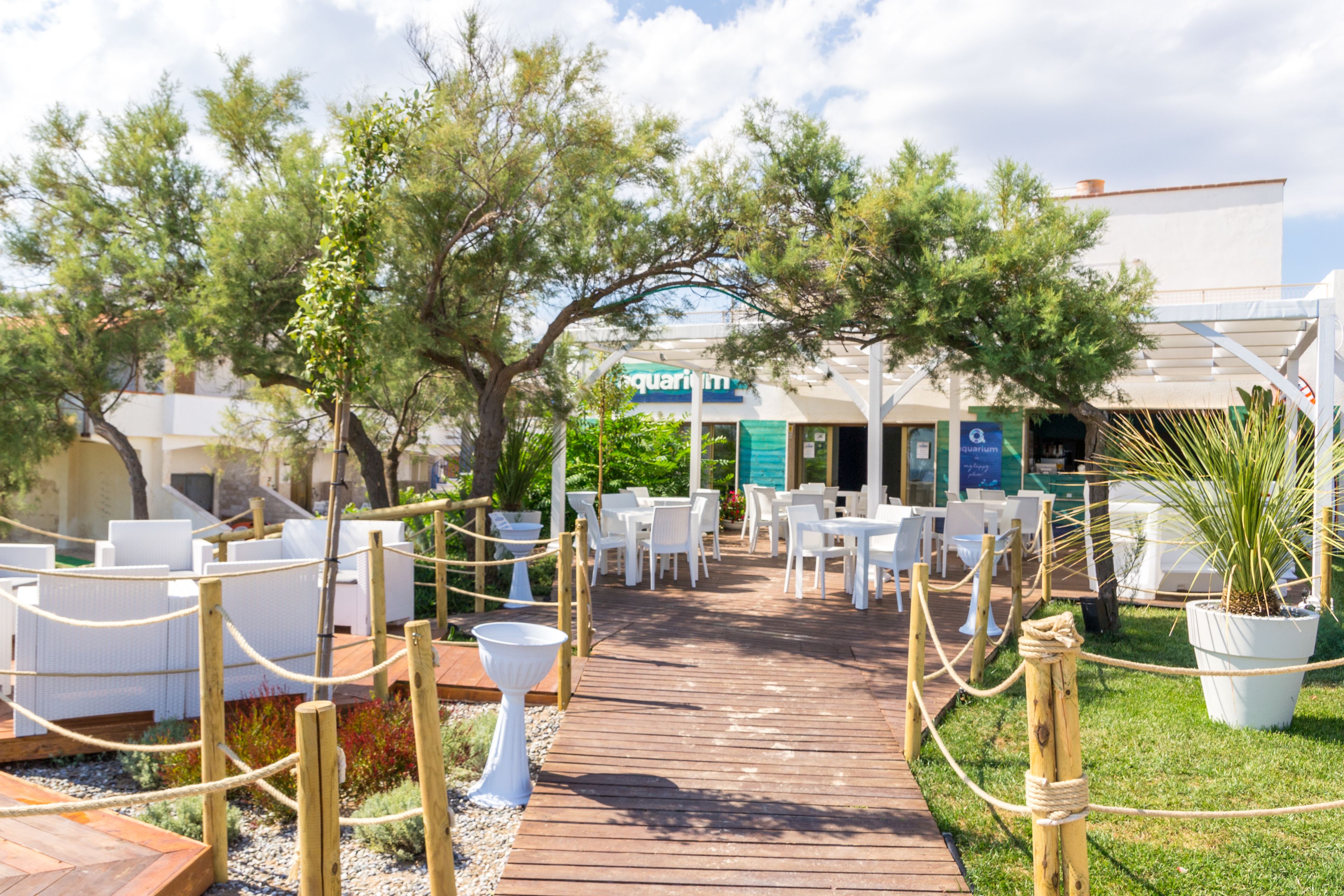 A wooden deck with white outdoor seating, surrounded by trees and potted plants, leading to a building with a sign that reads "aquarium".