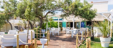 Une terrasse en bois avec des meubles de jardin blancs, entourée d’arbres et de plantes en pot, mène à un bâtiment sur lequel on peut lire l’inscription « aquarium ».