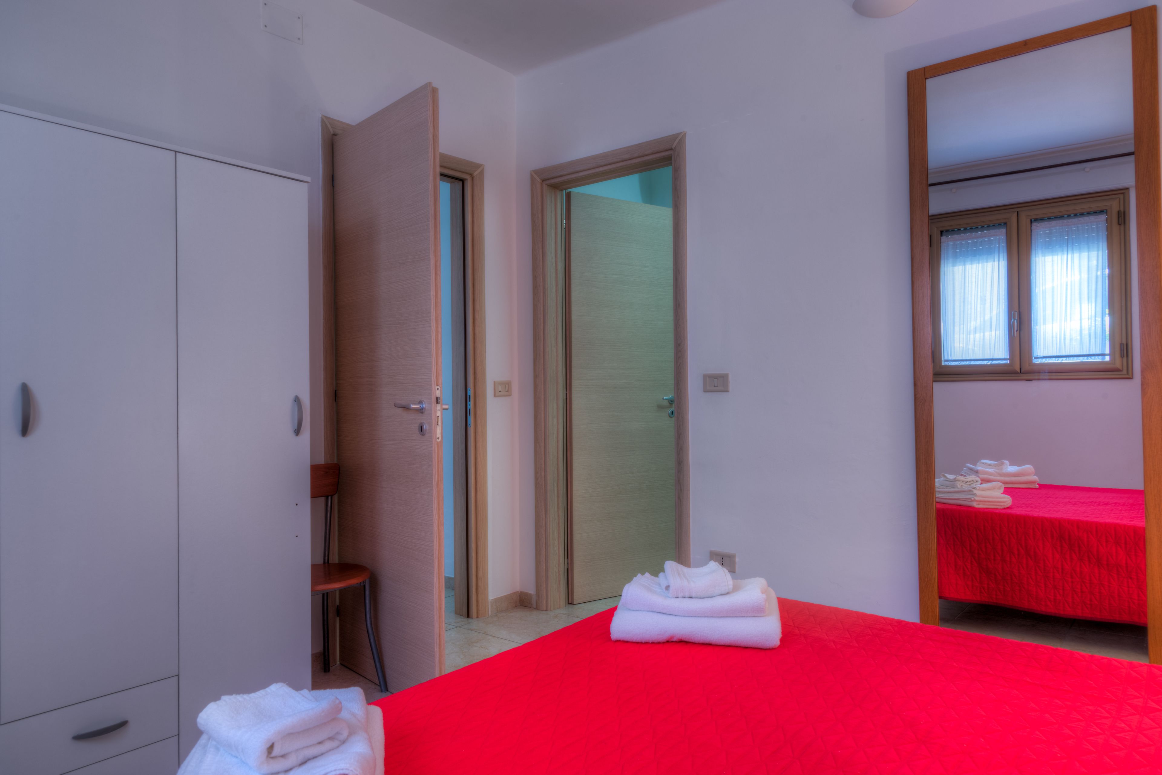 A hotel room with a red bedspread, a wooden wardrobe, a chair, and a small table with towels.