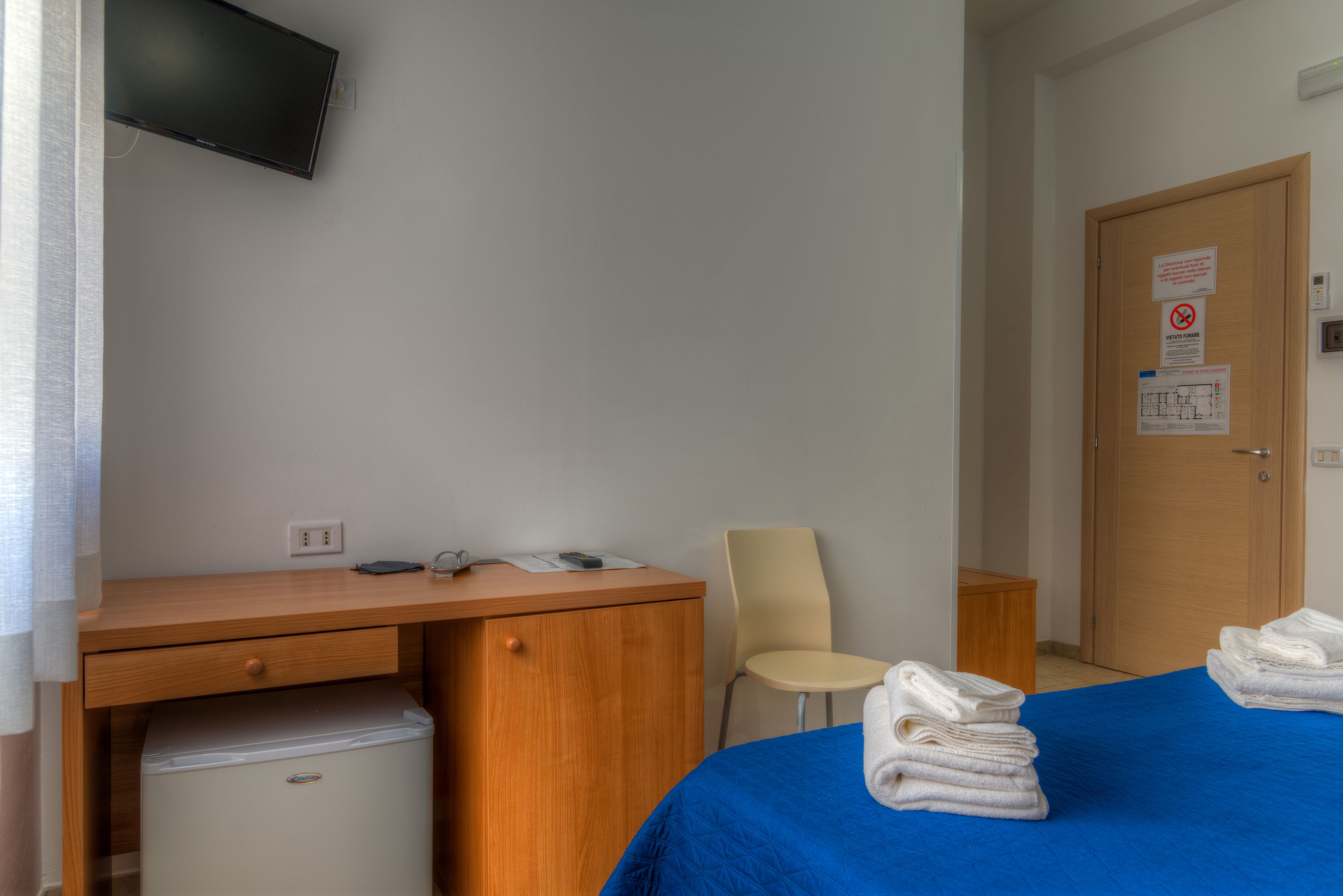 Habitación de hotel con cama, escritorio, silla, televisión y puerta con letrero.