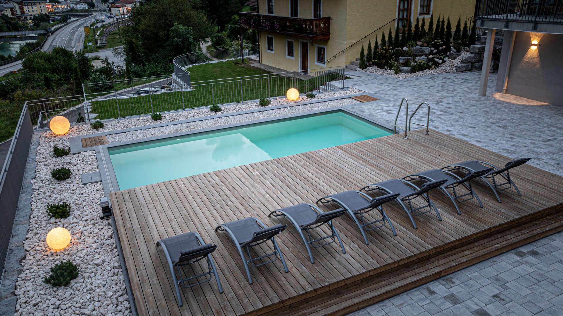 Una piscina con muelle de madera y sillones de descanso, rodeada de un jardín ajardinado y vistas a un pueblo.