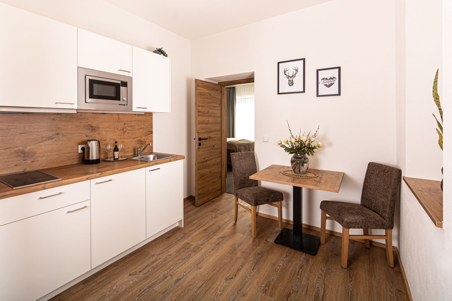 A compact kitchen with white cabinets, a wooden countertop, a microwave, and a sink. There is a dining table with two chairs and a vase with flowers.