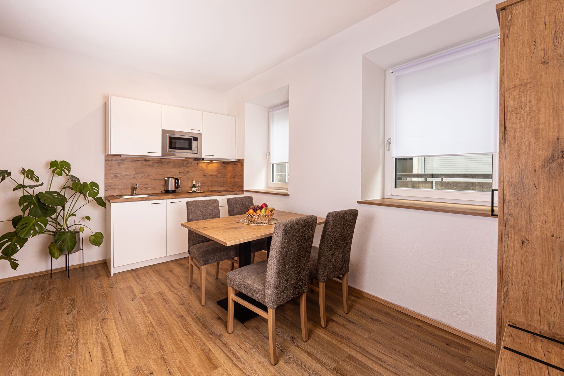 Une cuisine compacte avec des meubles blancs, un four à micro-ondes et un plan de travail en bois. On y trouve une table à manger avec quatre chaises et une plante en pot.
