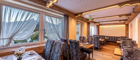 Un restaurant avec des tables en bois et des chaises à motifs, de grandes fenêtres avec des rideaux et un éclairage intérieur chaleureux.
