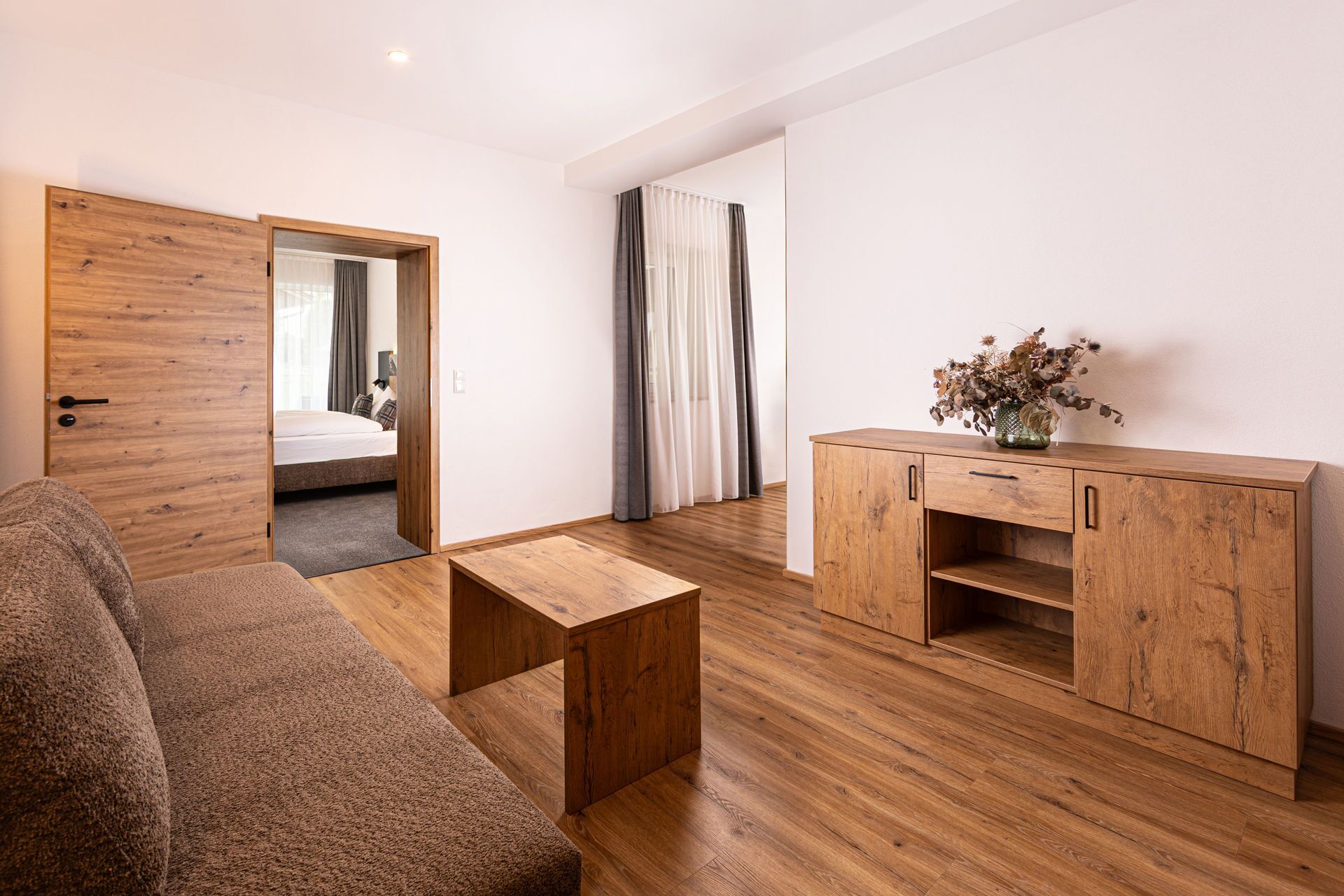 A modern hotel room with a wooden floor, a sofa, a small wooden table, a cabinet, and a bed with curtains.