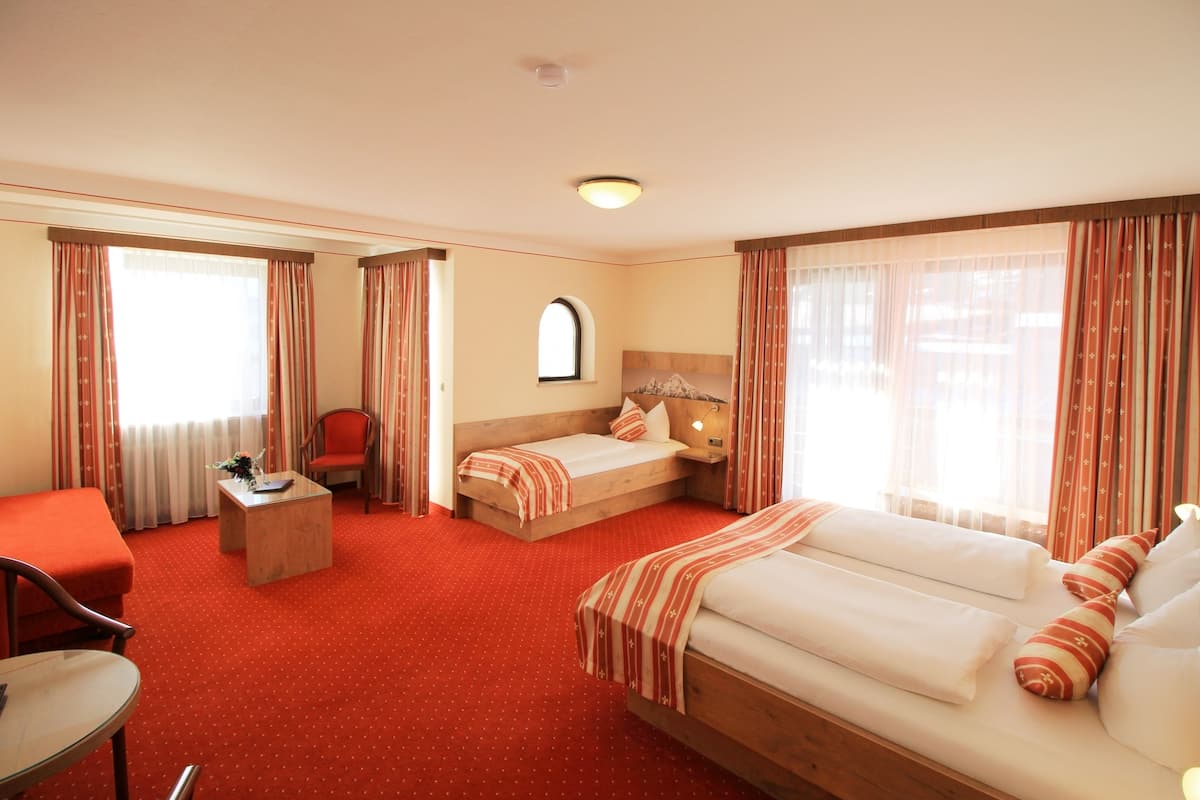 A hotel room with a red carpet, a bed, a sofa, a chair, a small table, and a window with curtains.