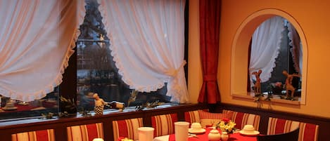 A dining area with a red tablecloth, white table settings, and a Christmas tree visible through the window.