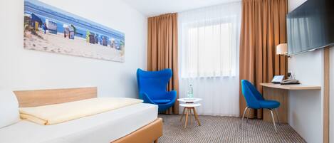 A hotel room with a bed, two blue chairs, a small table with a bottle, a TV on a stand, and a painting of a beach scene.