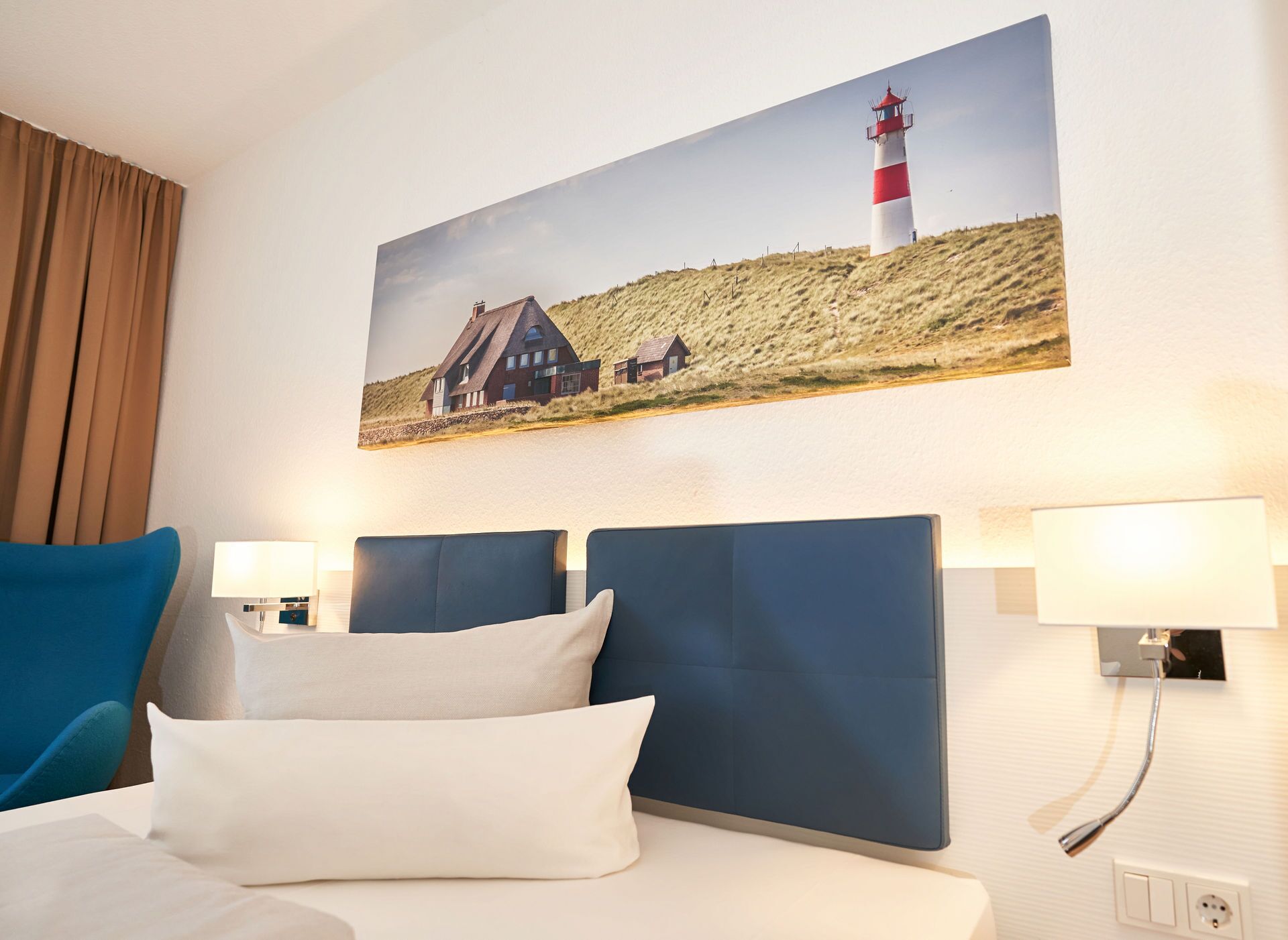 Habitación de hotel con cama, dos cabeceros, mesita de noche con lámpara y un cuadro en la pared de un faro y casas.