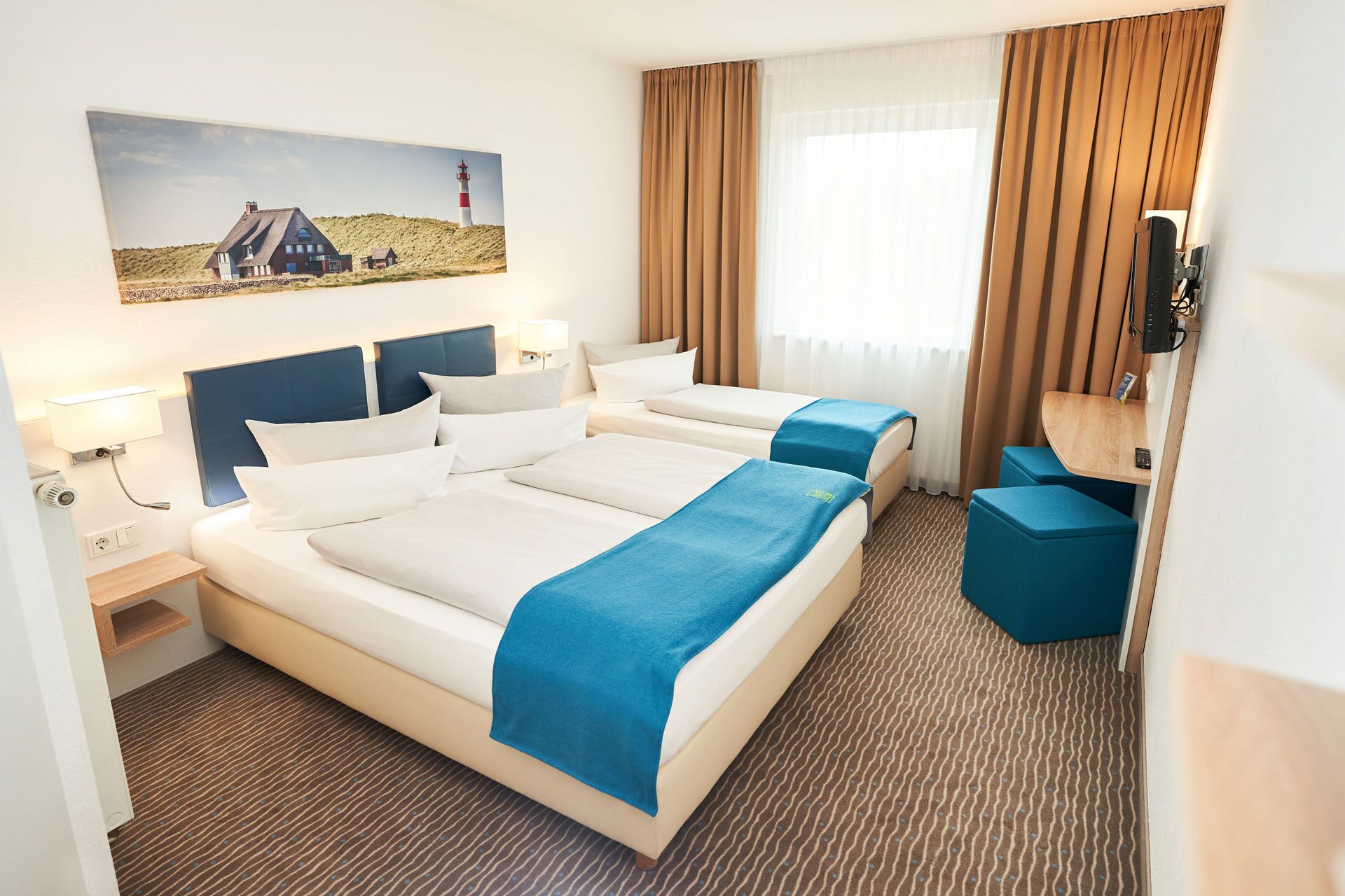 Quarto de hotel com uma cama grande, uma escrivaninha e uma cadeira. Há um quadro na parede com uma casa e um farol.
