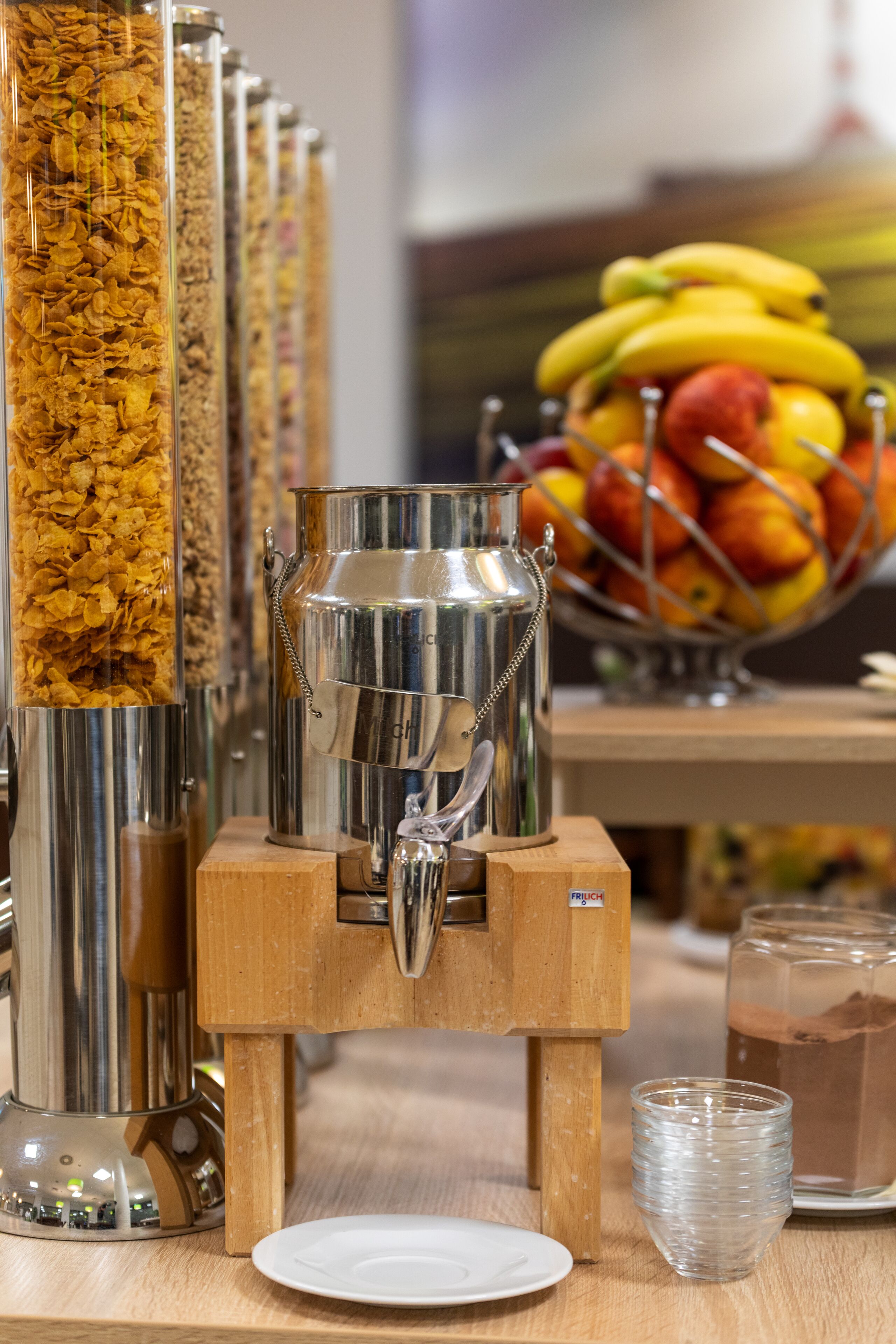 A beverage dispenser with a metal container and a wooden base, surrounded by glass containers of cereal.