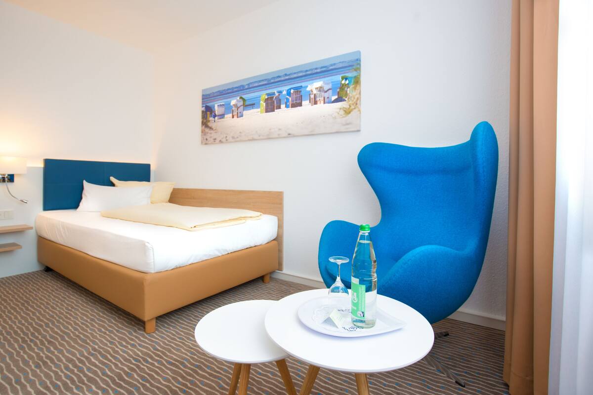 A hotel room with a bed, a blue armchair, a small table with a bottle and glass, and a painting of a beach scene on the wall.