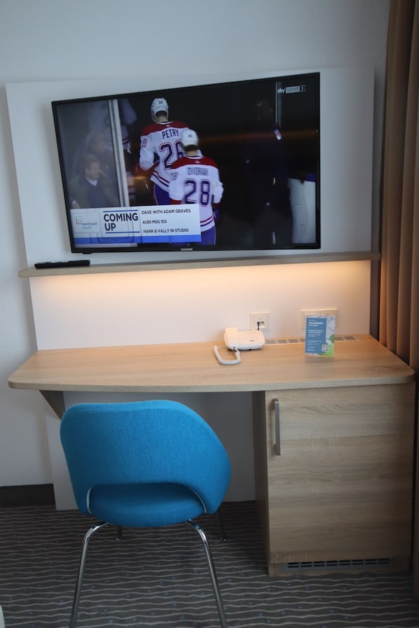 A hotel room with a flat-screen TV mounted on the wall, a wooden desk, a blue chair, a telephone, and a sign indicating a nearby hotel. - Hotel NordRaum (Bremen)