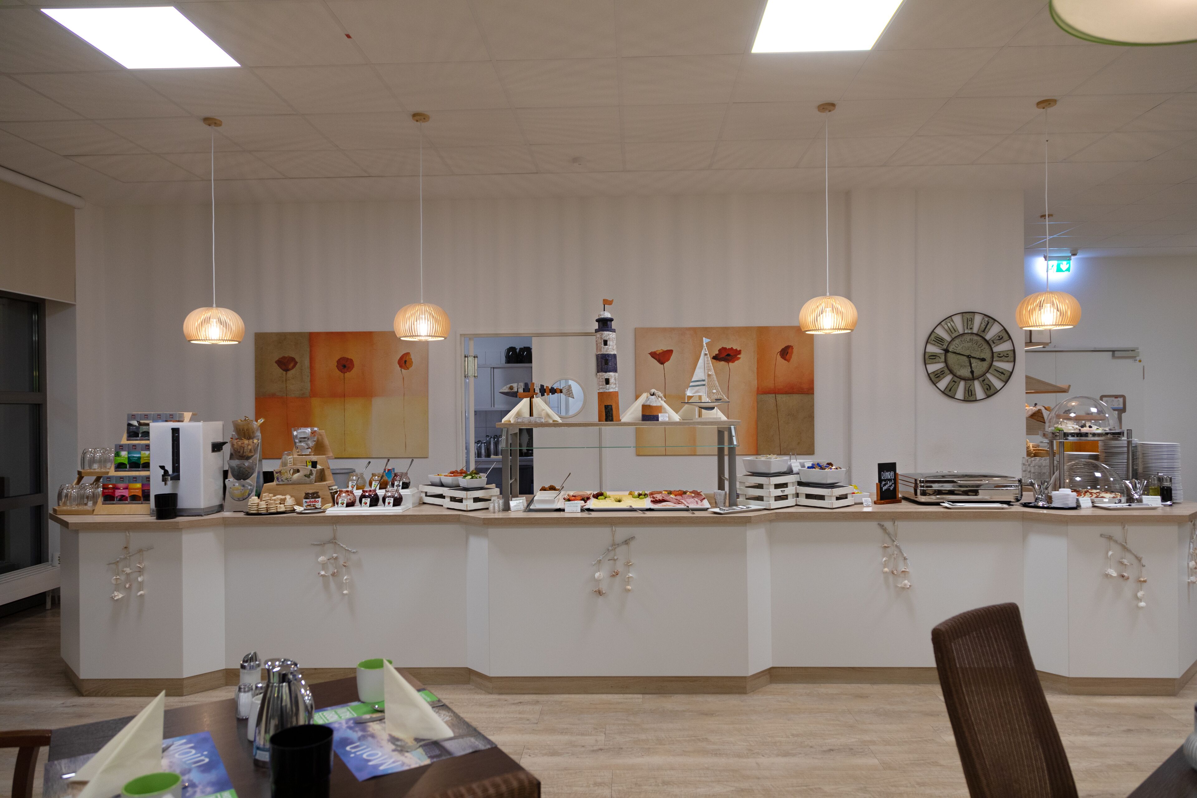 Una barra de desayuno con un reloj, lámparas colgantes y una pared decorativa con pinturas florales.