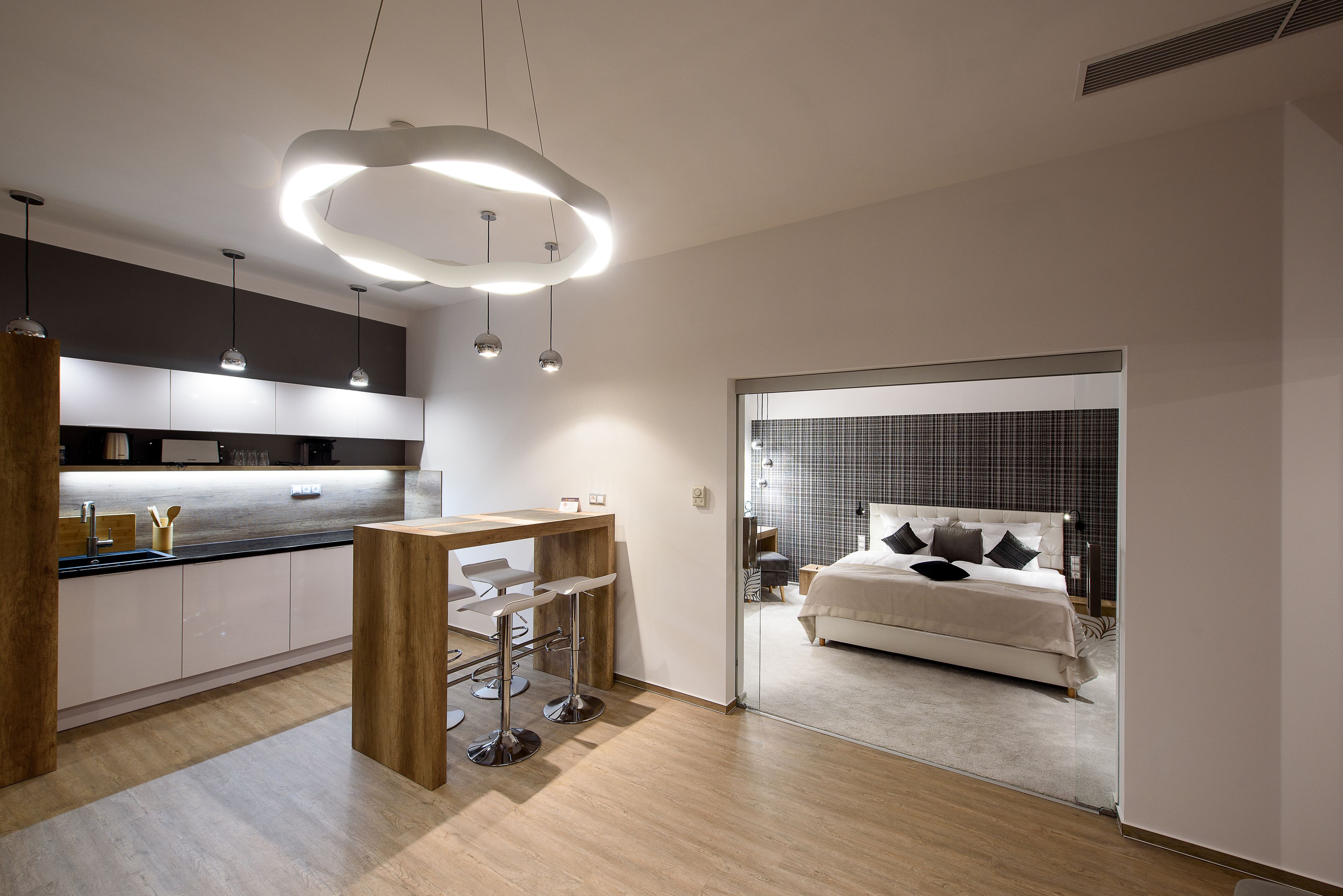 A modern hotel room with a kitchen area, a dining table, and a large bed.