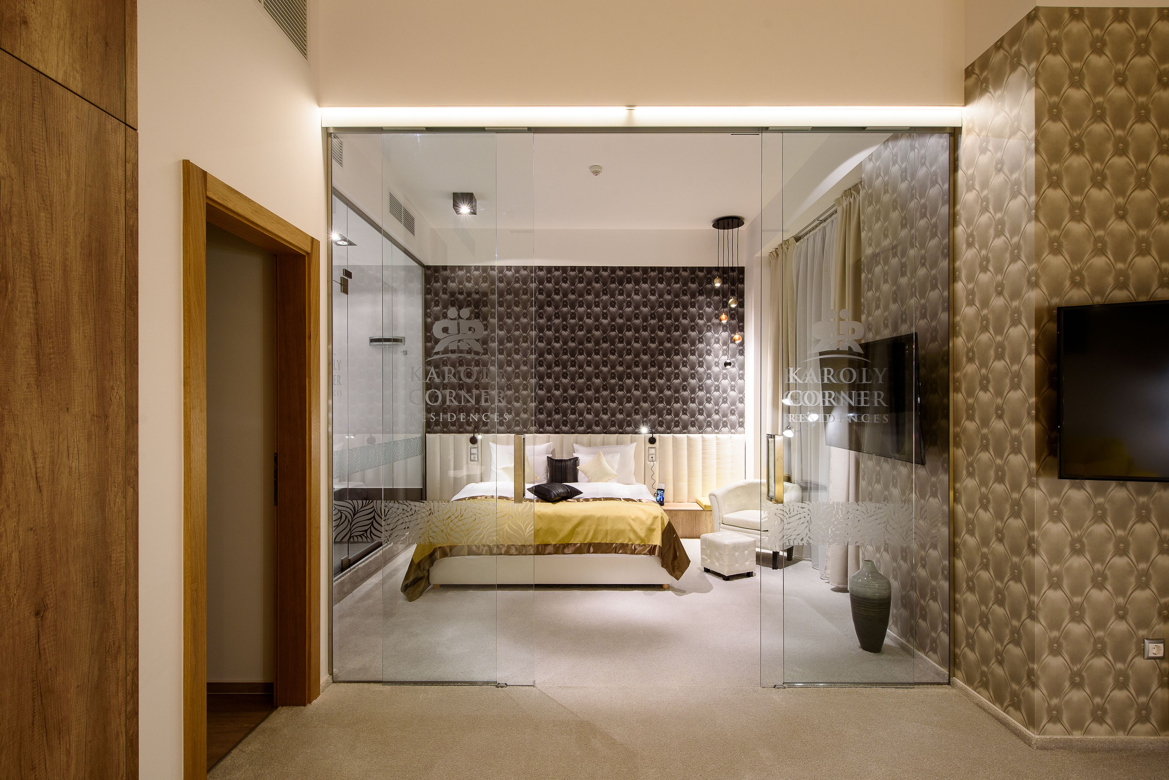 A modern bedroom with a glass partition, a bed with a patterned headboard, a flat-screen TV, and a large vase.