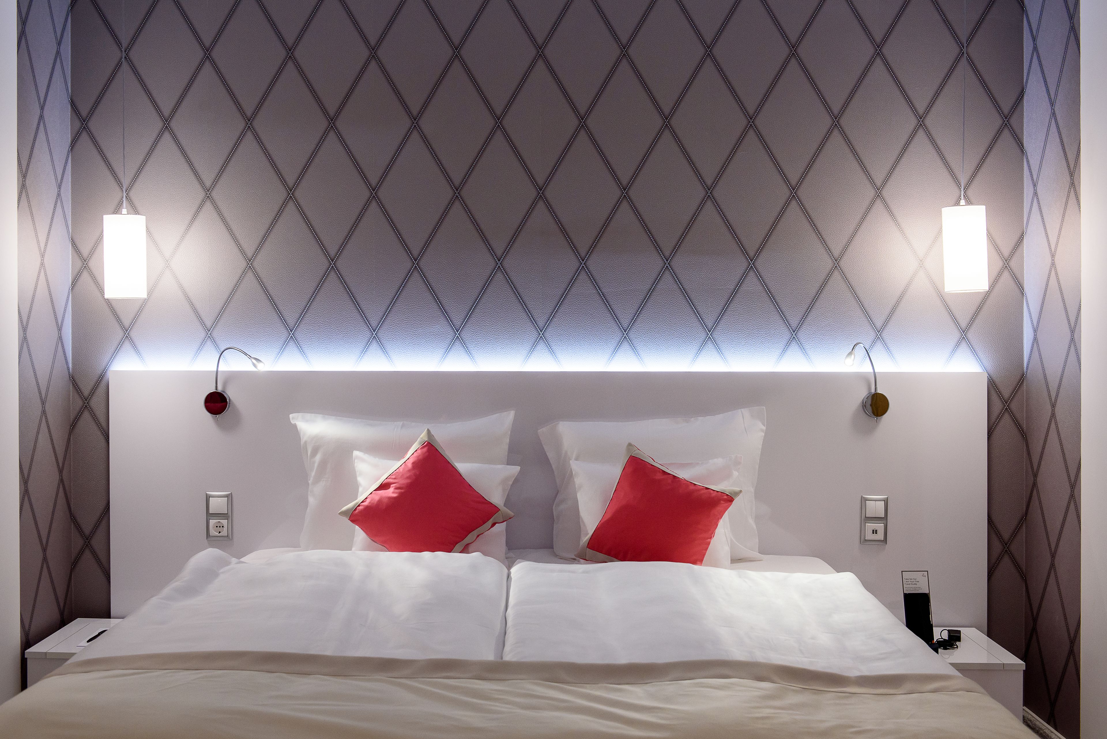 Una cama bien hecha con ropa blanca y dos almohadas rojas, una pared con patrón de rombos y dos lámparas de noche.