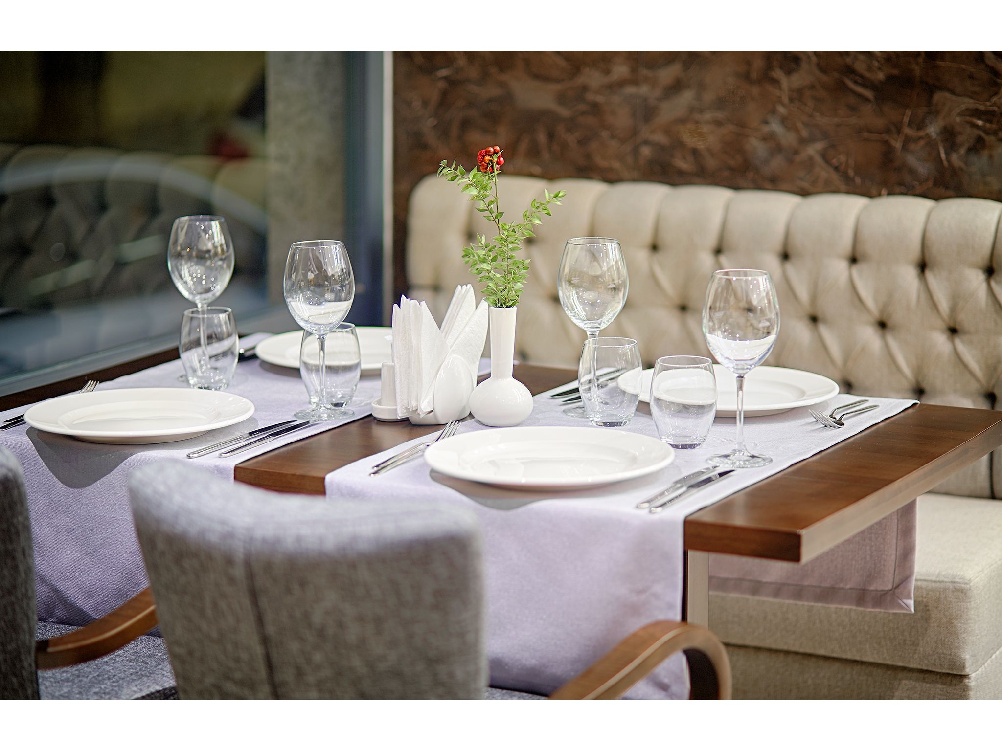 A dining table set with white plates, glasses, and cutlery, a vase with a plant, and a cushioned sofa in the background.