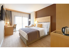 Quarto de hotel com cama, mesa de cabeceira, luminária, cadeira e vista da cidade através de grandes janelas.