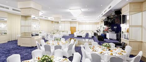 A banquet hall with round tables set for dining, white chairs, and a stage with musical equipment.