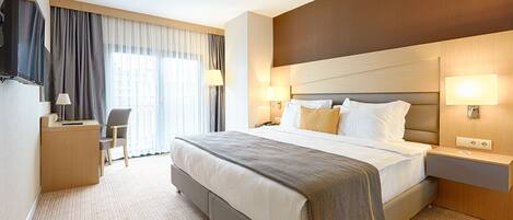 A hotel room with a large bed, bedside lamps, a desk with a chair, and a view of the city through the window.