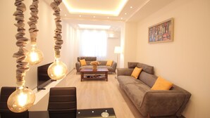 Uma sala de estar moderna com sofá, mesa de centro e poltronas. Apresenta luminárias pendentes e um quadro na parede.