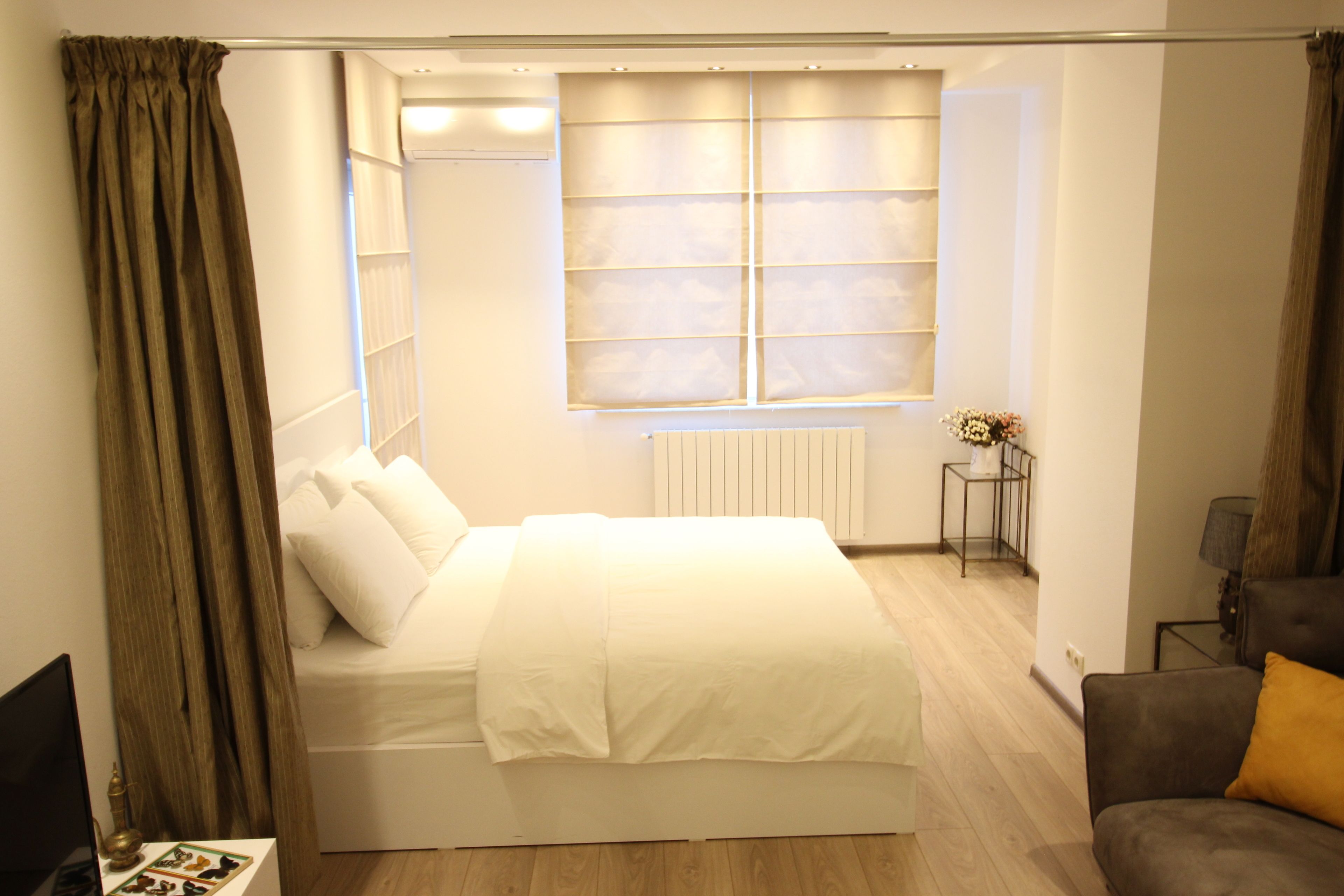 Un dormitorio con una cama grande, una mesita de noche, una ventana con persianas, un radiador y una pequeña mesa con lámpara.