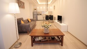 Uma sala de estar moderna com um sofá, mesa de centro e televisão. Uma cozinha é visível ao fundo.