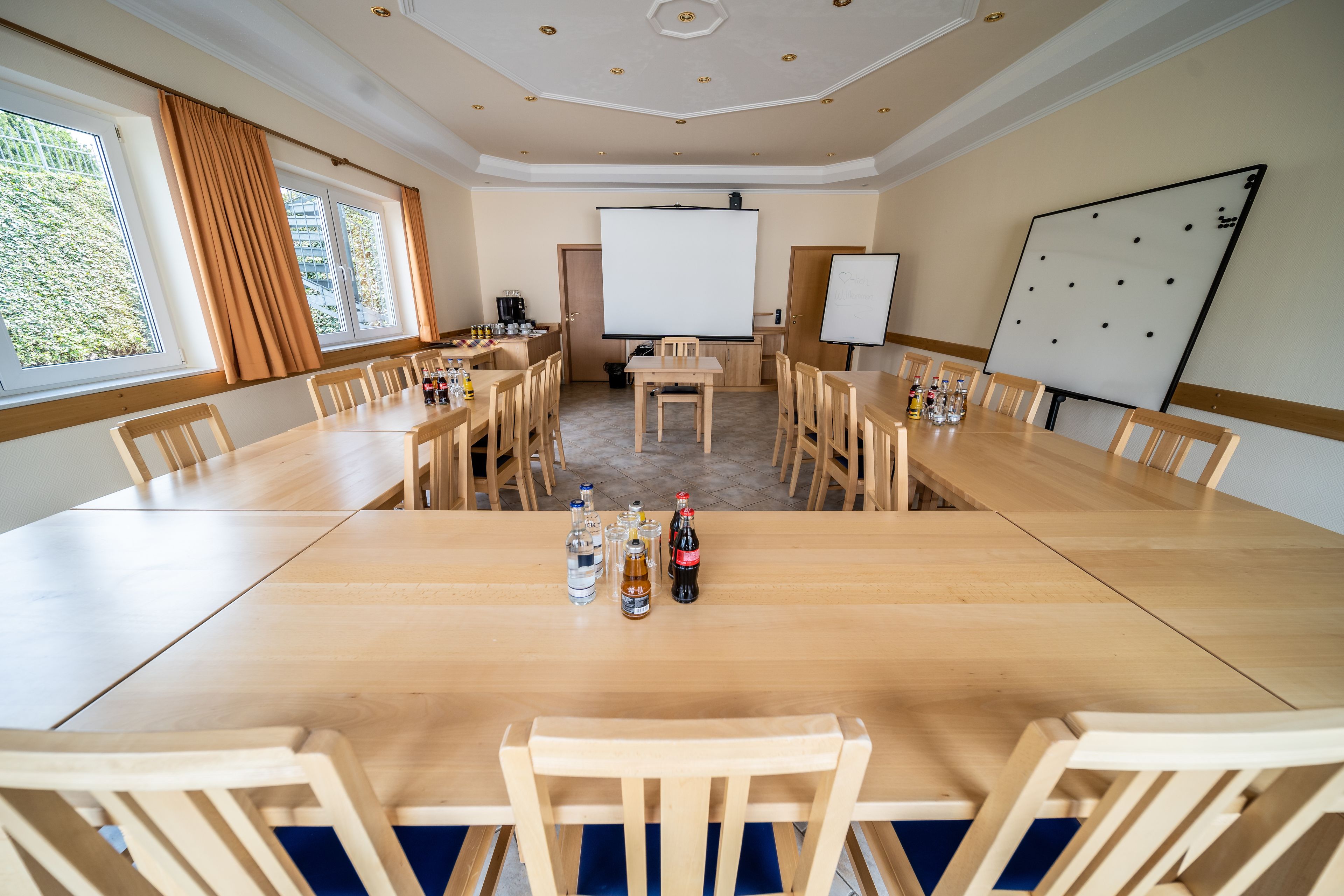 A conference room with a large wooden table, chairs, a projector screen, a whiteboard, and a window with curtains.