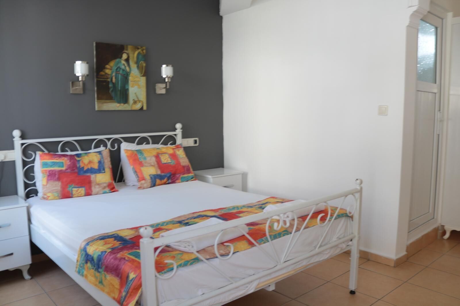 Ein Schlafzimmer mit einem weißen Metallbett, farbenfroher Bettwäsche, einem Nachttisch und einer Wandleuchte.