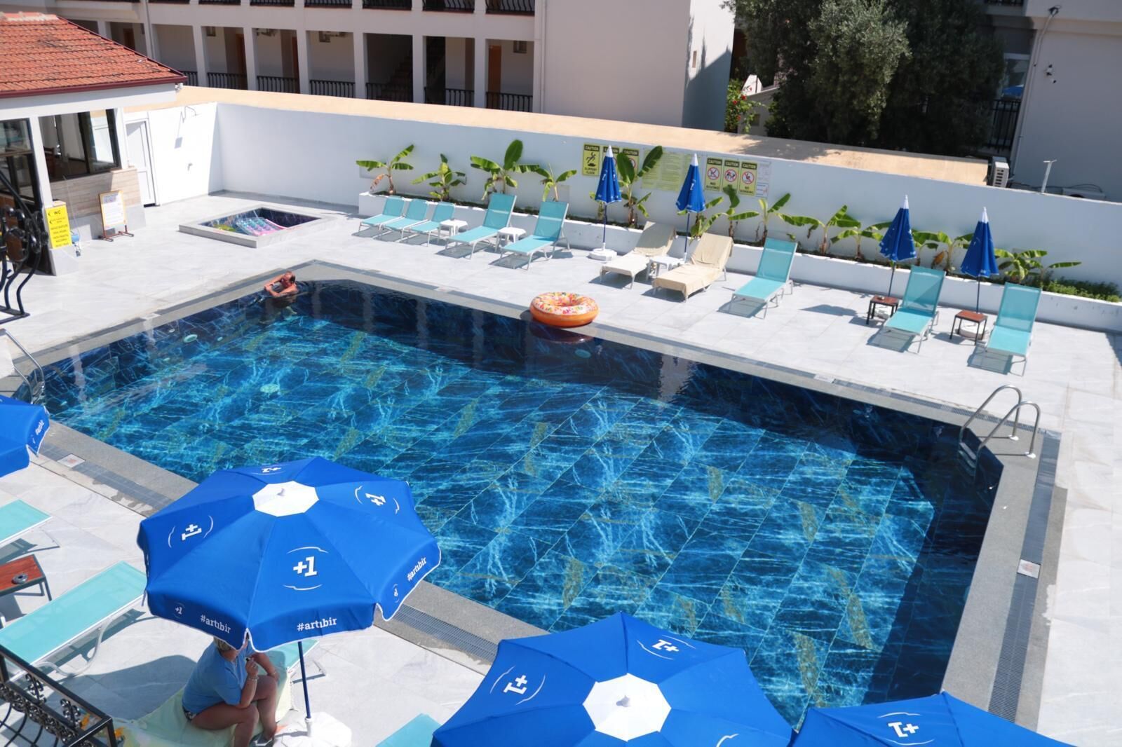 Una piscina con sillones de descanso y sombrillas, rodeada por un edificio blanco con balcones.