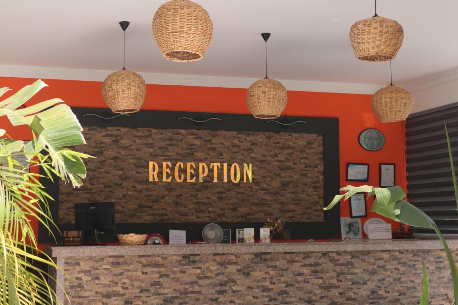 Zona de recepción con pared de ladrillo, un cartel que dice 'RECEPTION', lámparas colgantes de mimbre y una planta.