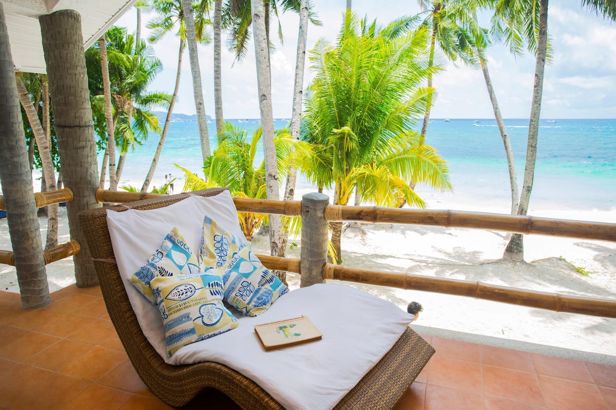 Silla de mimbre con cojín blanco y almohadones estampados, con vista a una playa con palmeras y el océano.