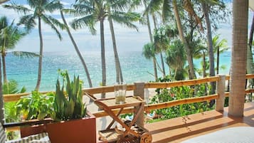 A balcony with a view of the ocean, palm trees, and a wooden cart with a glass and candle.