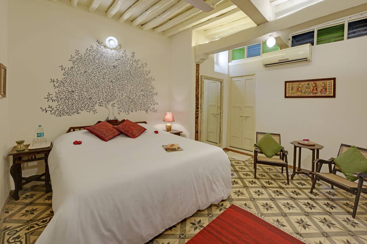A bedroom with a large bed, a wall mural, a small table with a lamp, and a seating area with chairs.