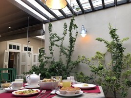 A breakfast table with various dishes, a teapot, and glasses, set in a room with a high ceiling and hanging plants.
