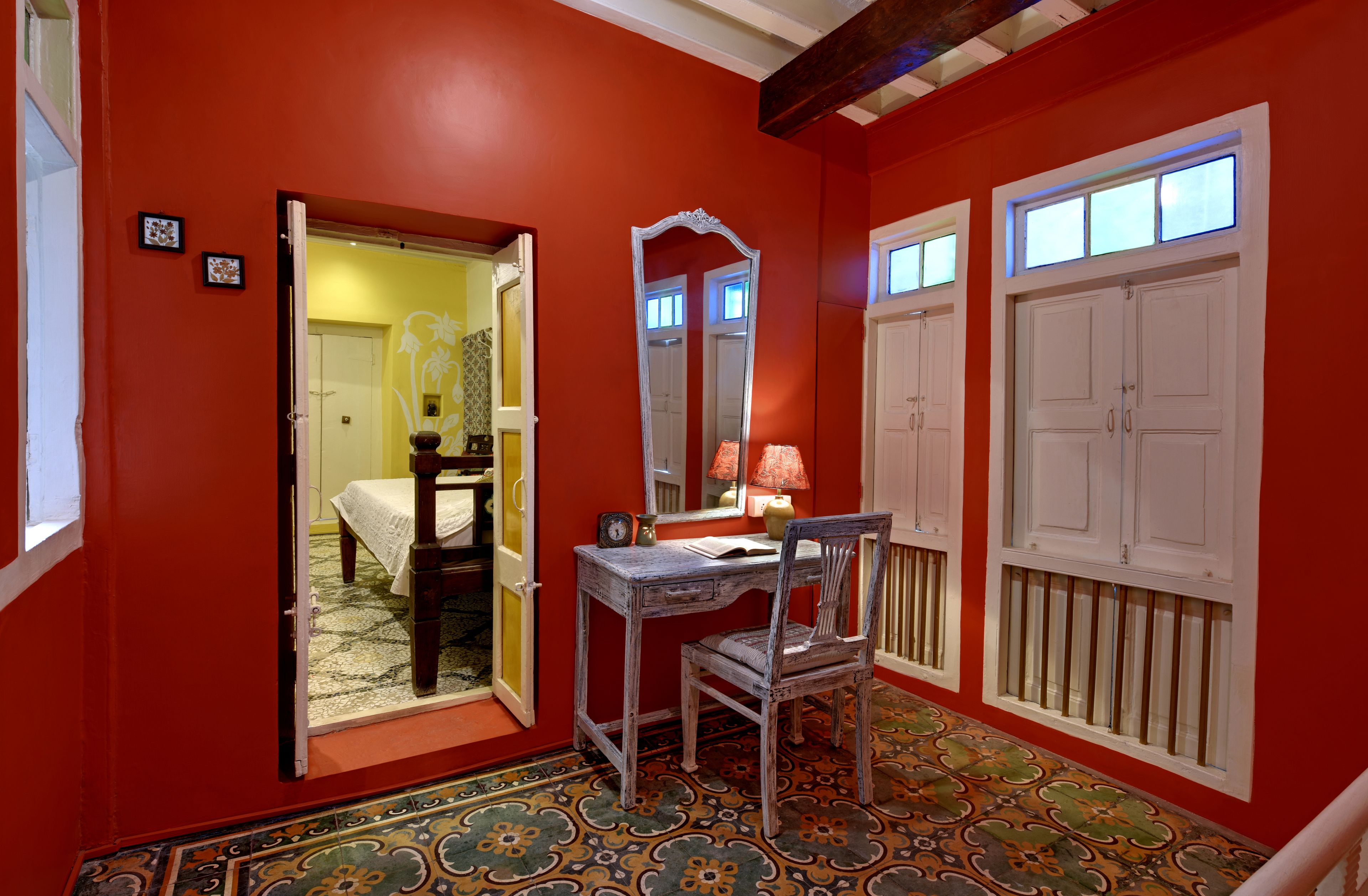 A room with a red wall, a wooden desk, a chair, a mirror, and a door leading to another room.