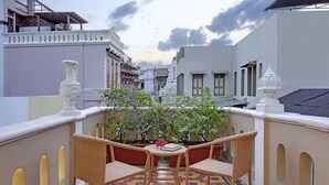 A balcony with wicker chairs and a small table, overlooking a garden with potted plants and a view of surrounding buildings. - French Haveli (Ahmedabad)