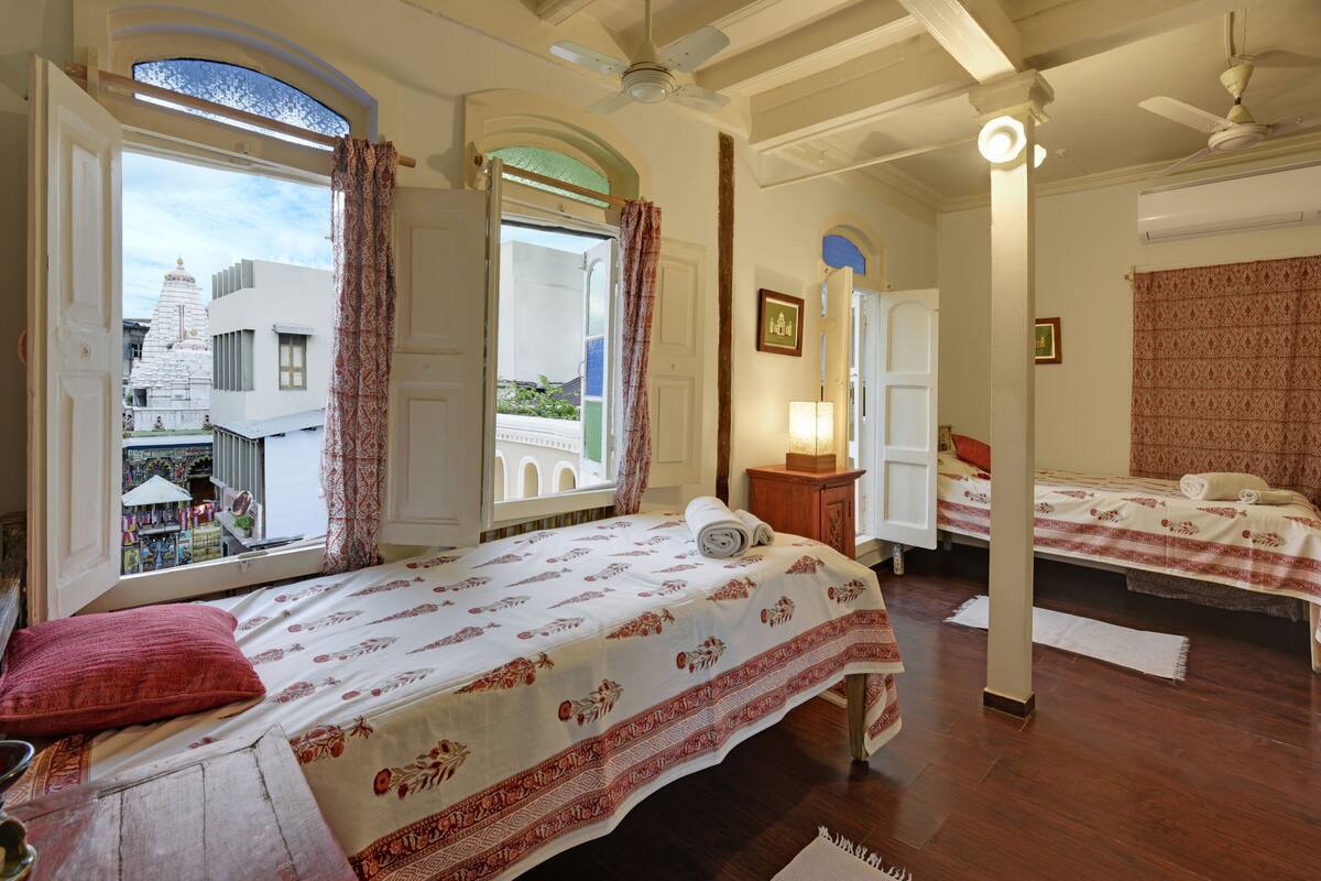 A hotel room with two beds, a large window with curtains, a ceiling fan, and a view of a temple outside.