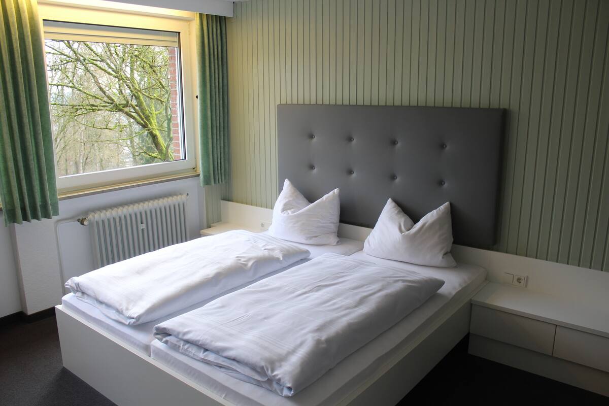 A twin bed with a grey headboard, white bedding, and a window with green curtains.