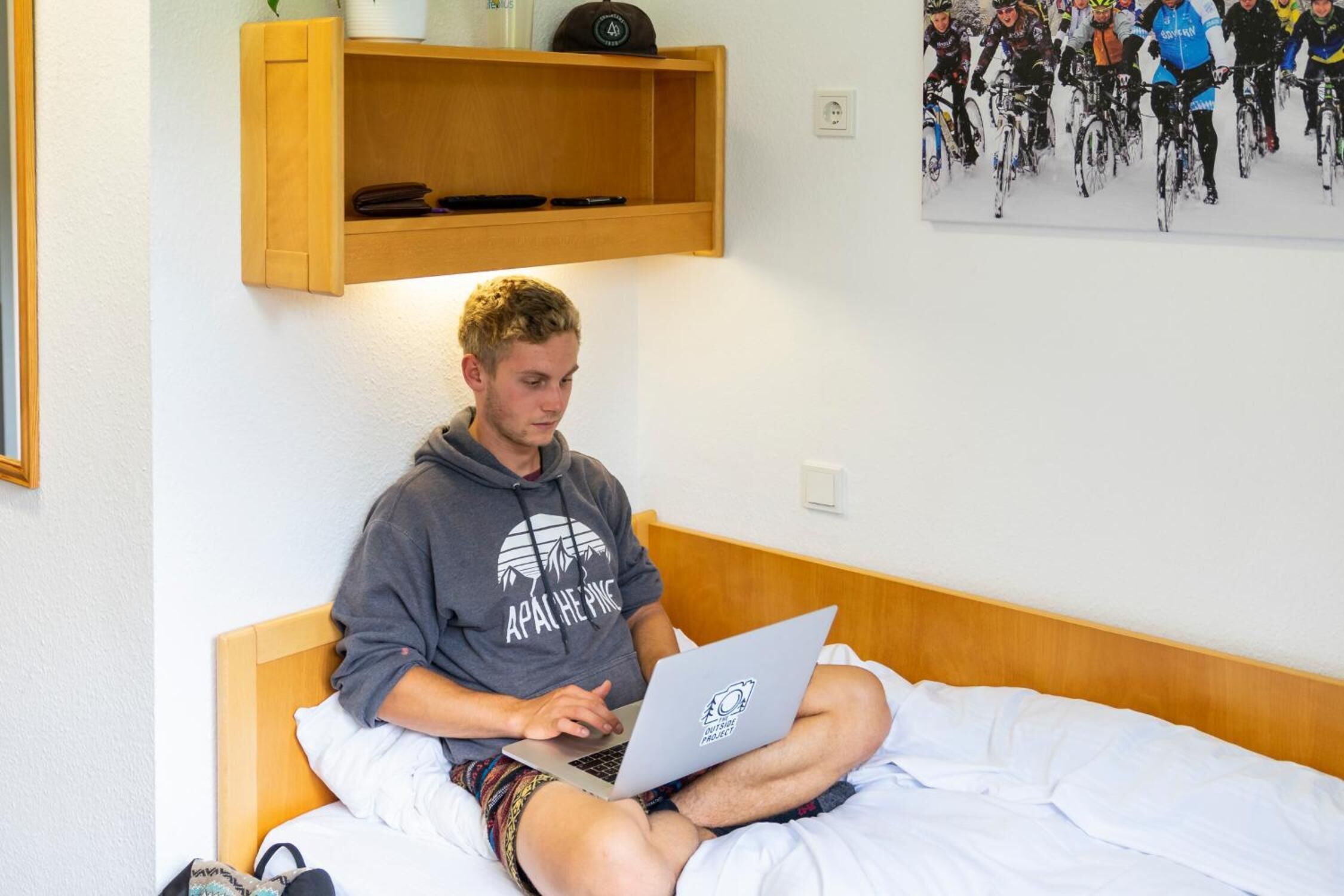 A person sitting on a bed with a laptop, a wooden shelf above, and a wall-mounted picture.