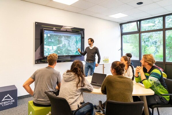 A presentation in a room with a large screen, a projector, and a group of people seated around a table. - Adventure Campus (Treuchtlingen)