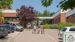 Un estacionamiento con varios coches y un grupo de personas caminando hacia un edificio con entrada de vidrio.