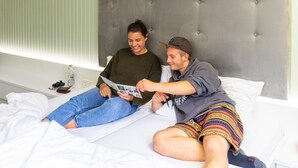 Una cama con cabecera capitonada, ropa de cama blanca y una mesita de noche con una lámpara y un objeto pequeño.